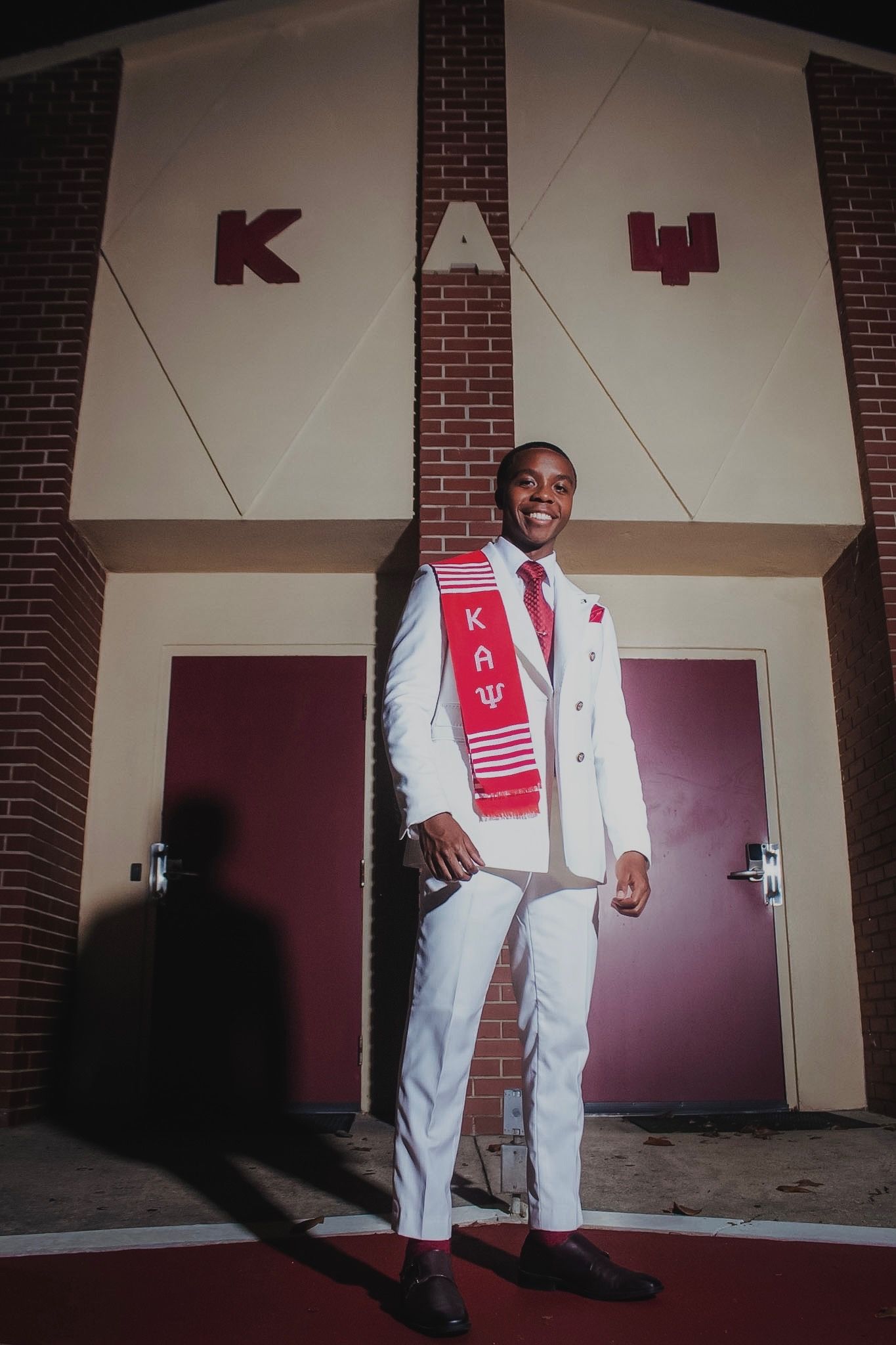 Sean Inman in a white suit and crimson kente stole, smiling outside the chapter house