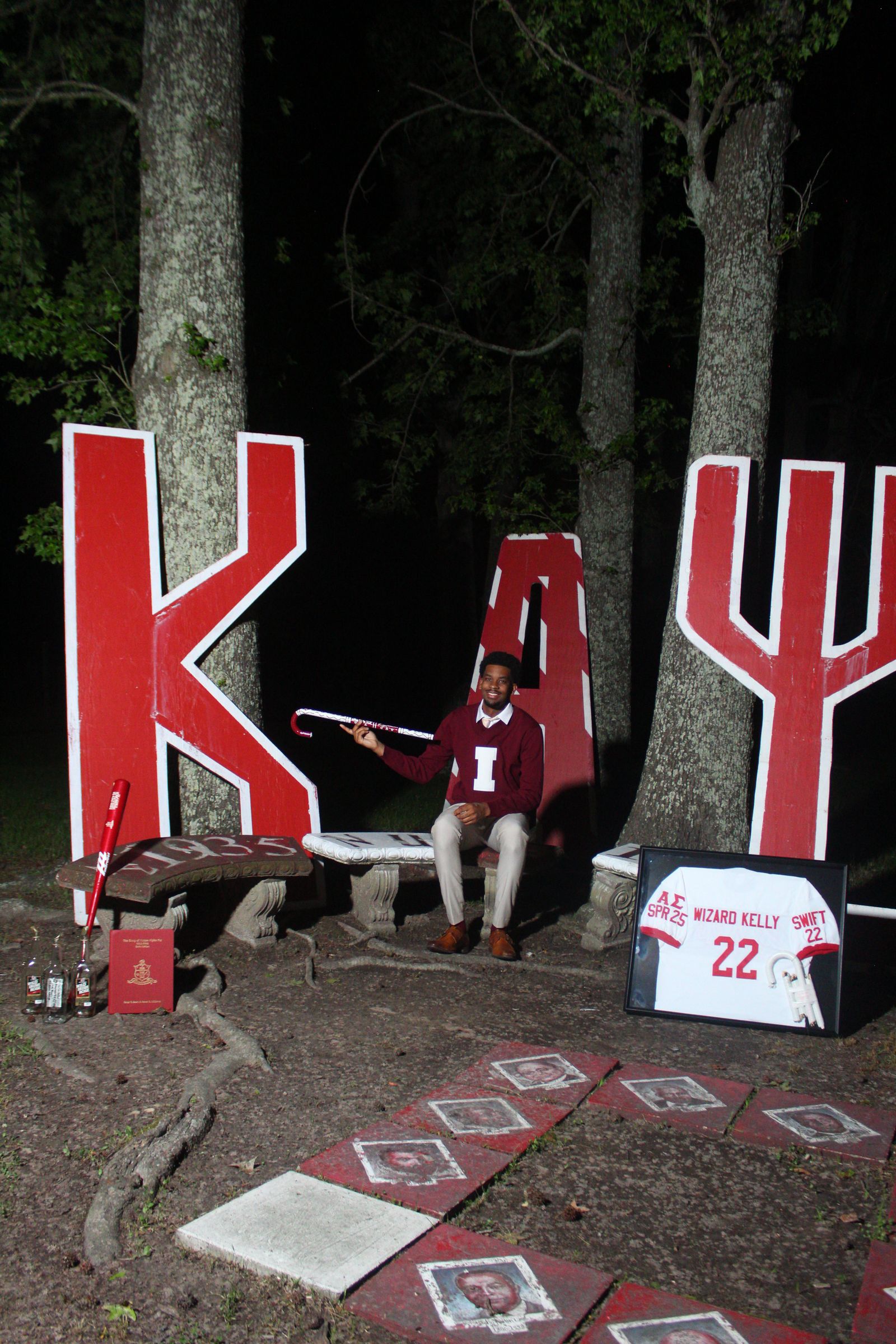 A brother seated among large crimson Greek letters in a wooded grove at night